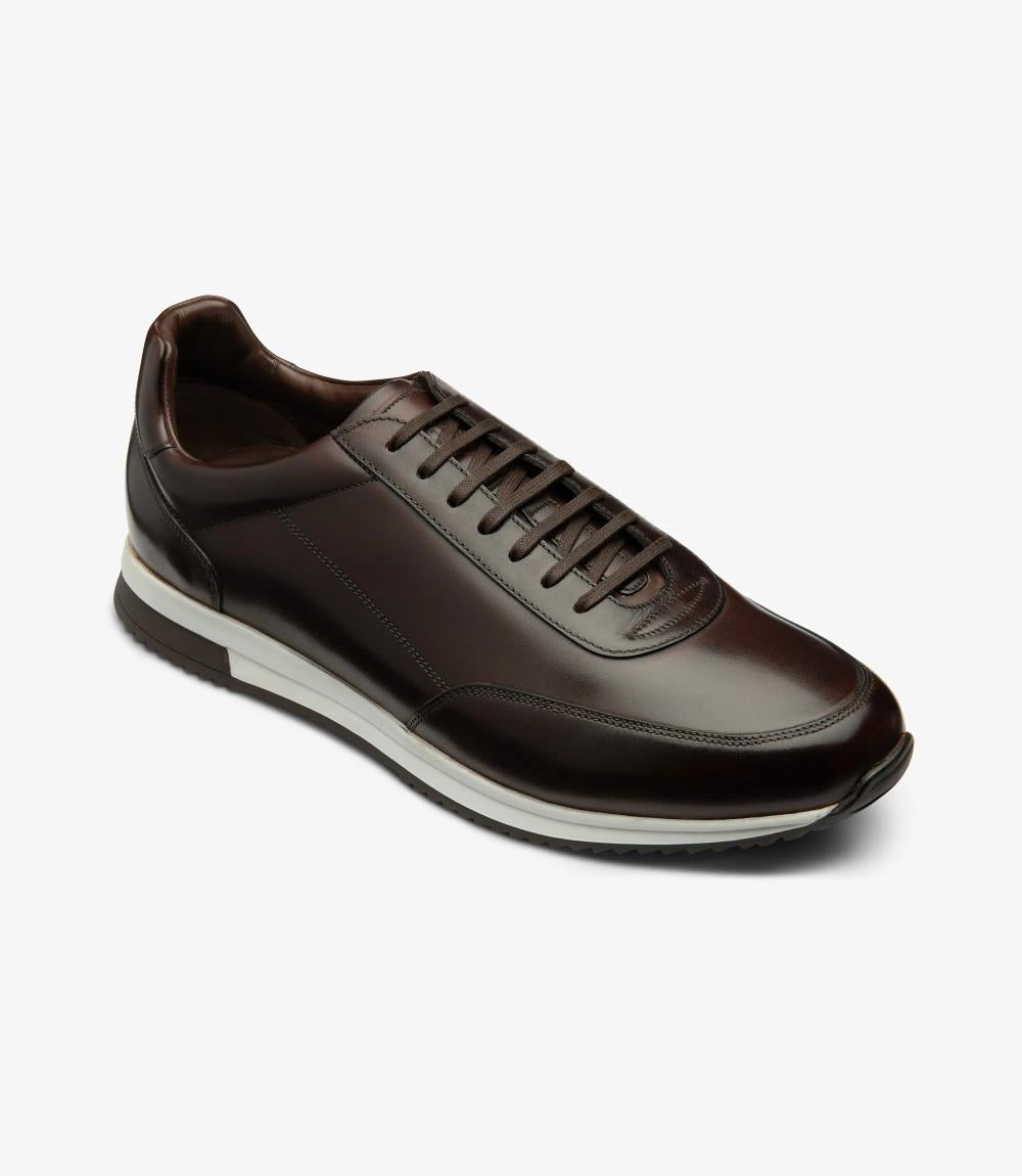 An angled shot capturing the Loake Bannister Dark Brown Leather Trainer from the side, emphasizing its elegant contours and bold toe shape. The high-quality craftsmanship and attention to detail are evident, making it a versatile choice for casual wear.