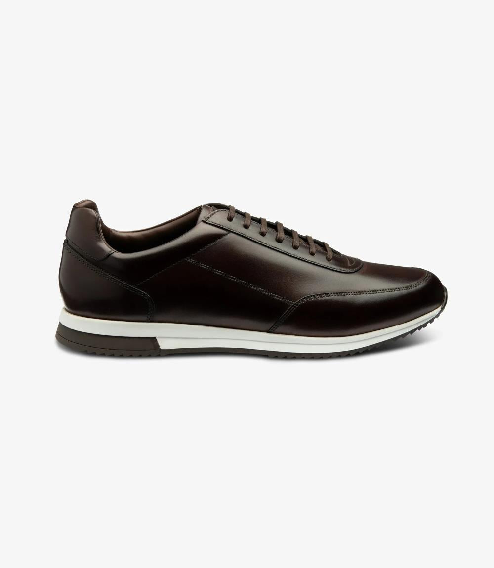 A side view showcasing the sleek silhouette of the Loake Bannister Dark Brown Leather Trainer, highlighting its refined design and premium materials. The rich cedar-burnished calf leather gleams under the light, while the cemented rubber sole provides durability and traction.