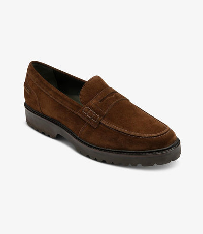 From a dynamic angle, the Loake Crux Brown Suede Loafer reveals its modern flair and impeccable craftsmanship. The rich brown suede leather gleams under the light, while the commando sole adds durability and grip. The sleek profile of the shoe highlights its versatility and sophistication.