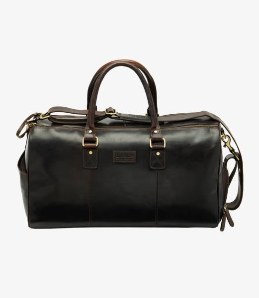 The Loake Devon Dark Brown Leather Overnight Bag is displayed from the front, showcasing its rich, vegetable tanned leather finish. The sleek, minimalist design is accentuated by the high-quality stitching and polished leather texture. The sturdy leather handles with a firm grip are prominently visible, emphasizing the bag's practicality and elegance.