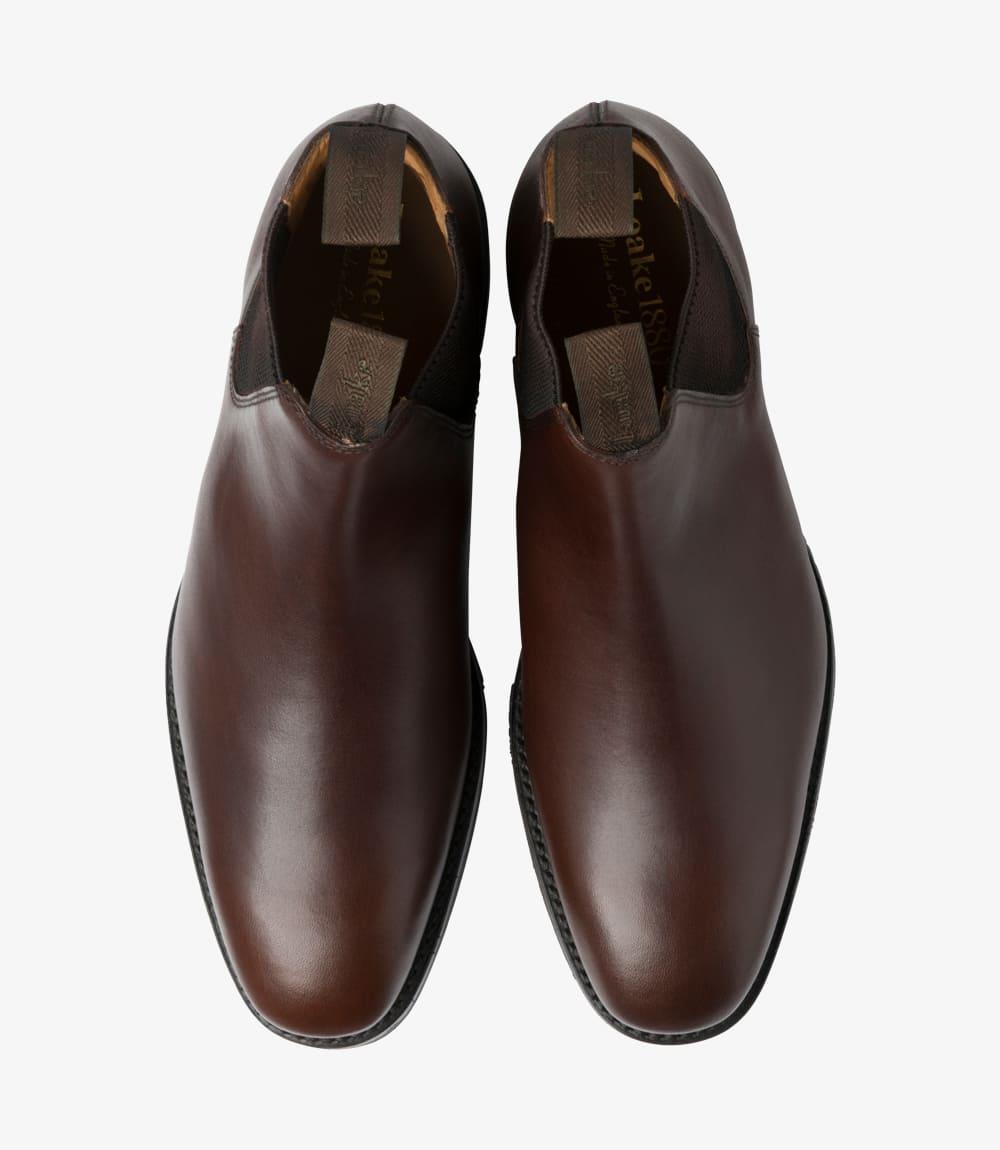 This overhead shot features both Chelsea boots, emphasizing their matching symmetry and design. The smooth surface of the dark brown waxed leather shines under the light, showcasing its premium quality. The elastic panels and ankle height are clearly visible, highlighting the Chelsea boot style's characteristic features. The boots sit neatly side by side, demonstrating their elegant profile and the craftsmanship involved in their creation.