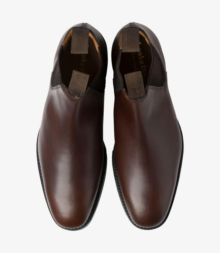 This overhead shot features both Chelsea boots, emphasizing their matching symmetry and design. The smooth surface of the dark brown waxed leather shines under the light, showcasing its premium quality. The elastic panels and ankle height are clearly visible, highlighting the Chelsea boot style's characteristic features. The boots sit neatly side by side, demonstrating their elegant profile and the craftsmanship involved in their creation.