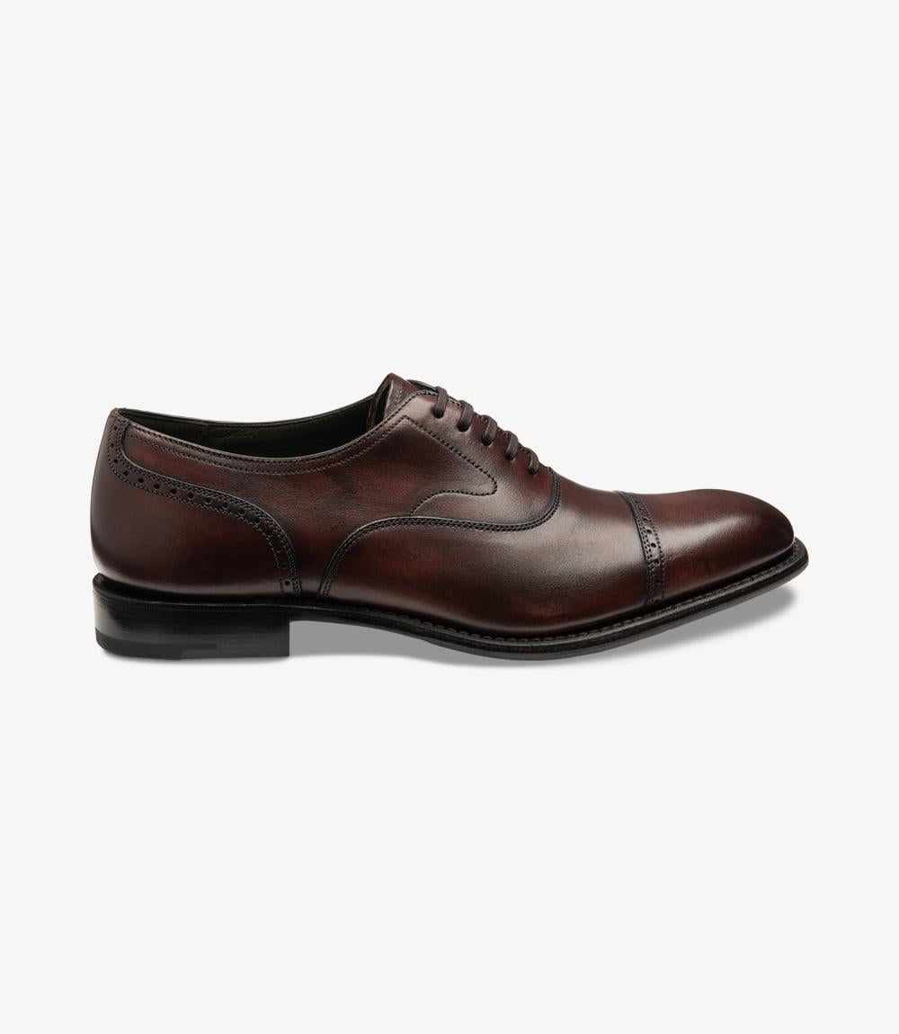 The shoe is displayed from the side, showcasing its elegant silhouette. The deep burgundy hand patina calf leather shines softly in the light, highlighting the subtle punched detailing along the straight toe-cap. The Goodyear welted leather and rubber sole is visible, emphasizing its durability and classic style. The shoe's sleek profile is enhanced by the bold toe shape, making it a sophisticated addition to any outfit.