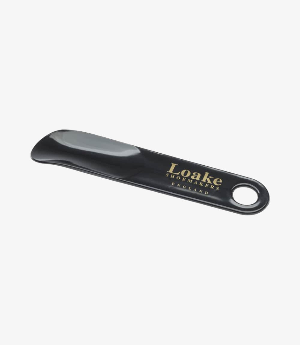 The Loake Black Plastic Shoe Horn is displayed in its closed position, showcasing its sleek and smooth design. The shoe horn features a shiny black finish, reflecting light subtly, while the Loake branding is prominently visible near the top. The gently curved shape of the horn tapers towards the bottom, emphasizing its ergonomic design, which facilitates easy shoe entry.